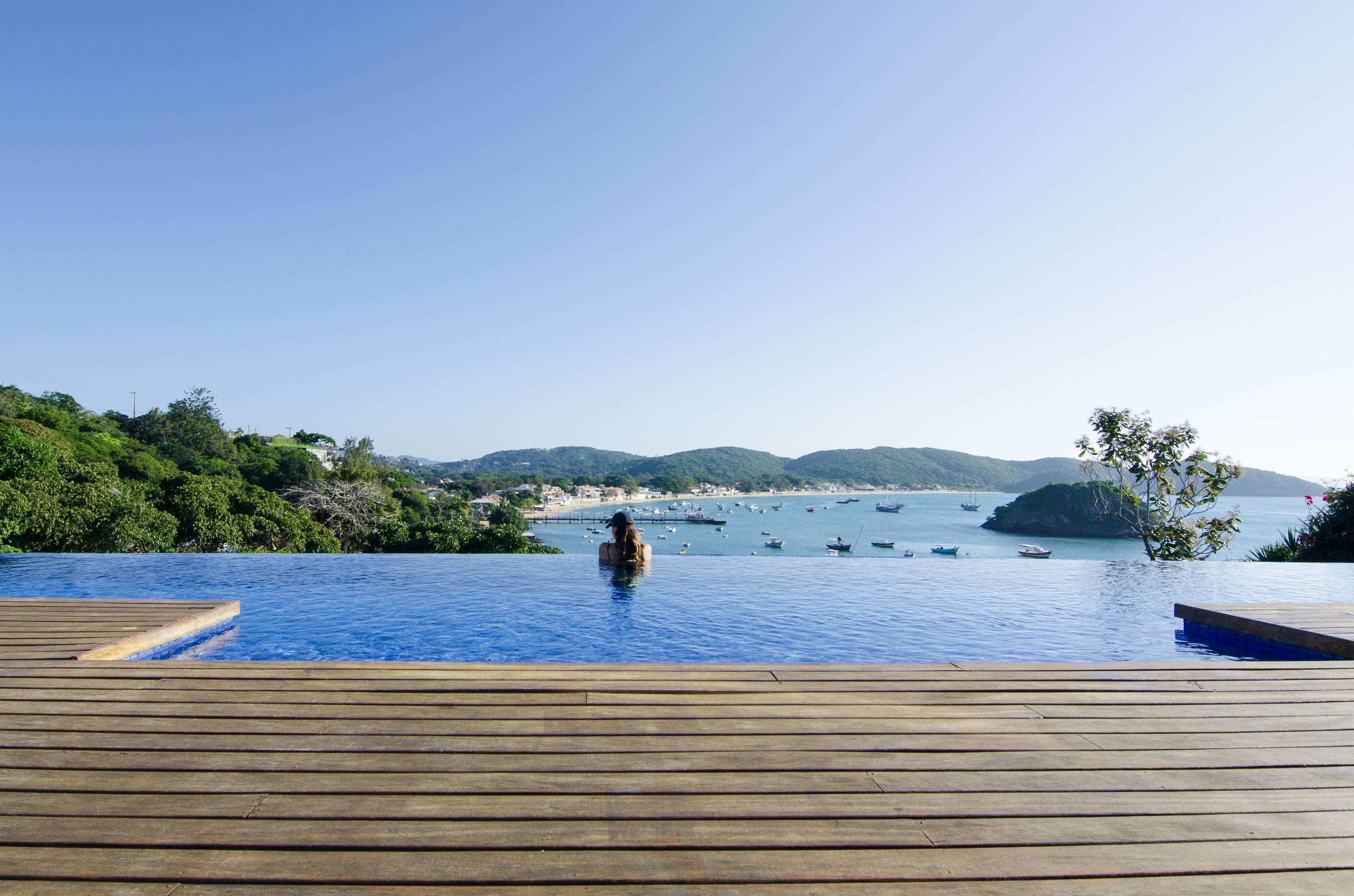 Una piscina infinita con una terraza de madera, con vistas a una bahía con barcos y una costa lejana.
