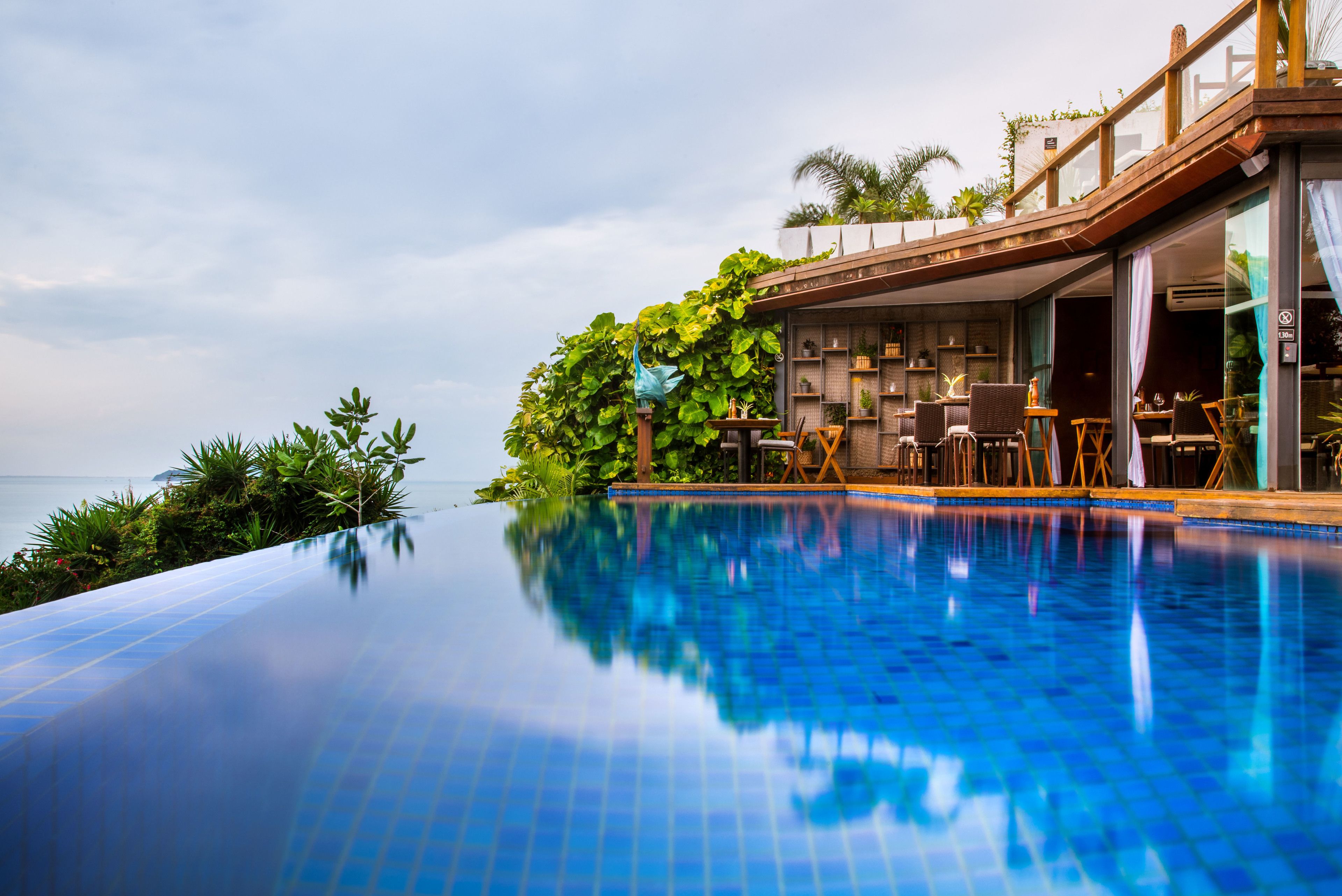 A infinity pool with a view of the ocean, surrounded by lush greenery and a wooden deck with outdoor seating.