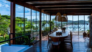 A wooden deck with a dining area, large windows offering a view of a beach and boats, and potted plants. - Abracadabra Boutique Hotel (Búzios)