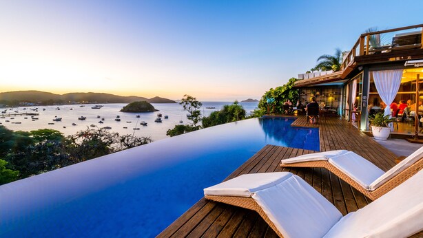 An infinity pool with wooden deck, lounge chairs, and a view of boats on the water. - Abracadabra Boutique Hotel (Búzios)