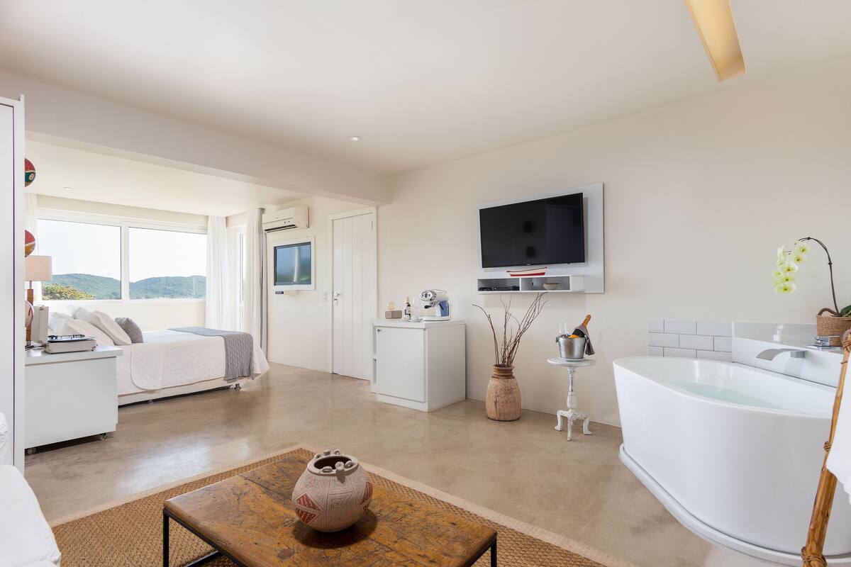 Una habitación de hotel moderna con un ventanal grande, un televisor de pantalla plana, una bañera blanca y un escritorio pequeño con un florero.