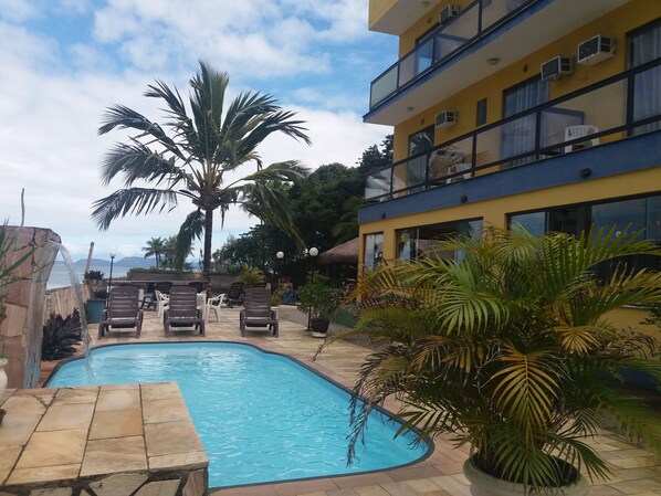 A swimming pool with lounge chairs, palm trees, and a building with balconies and air conditioning units. - Pousada dos Golfinhos (Angra dos Reis)