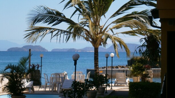Zona junto a la piscina con sillones de descanso, una palmera y vistas al mar y montañas distantes.