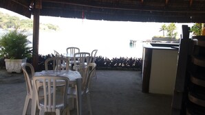 Un patio al aire libre con sillas de plástico y una mesa, un techo de paja y vista al mar.