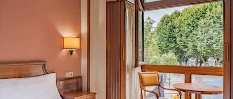 Habitación de hotel con cama, mesita de noche, una silla, una pequeña mesa y ventana con vista a árboles.