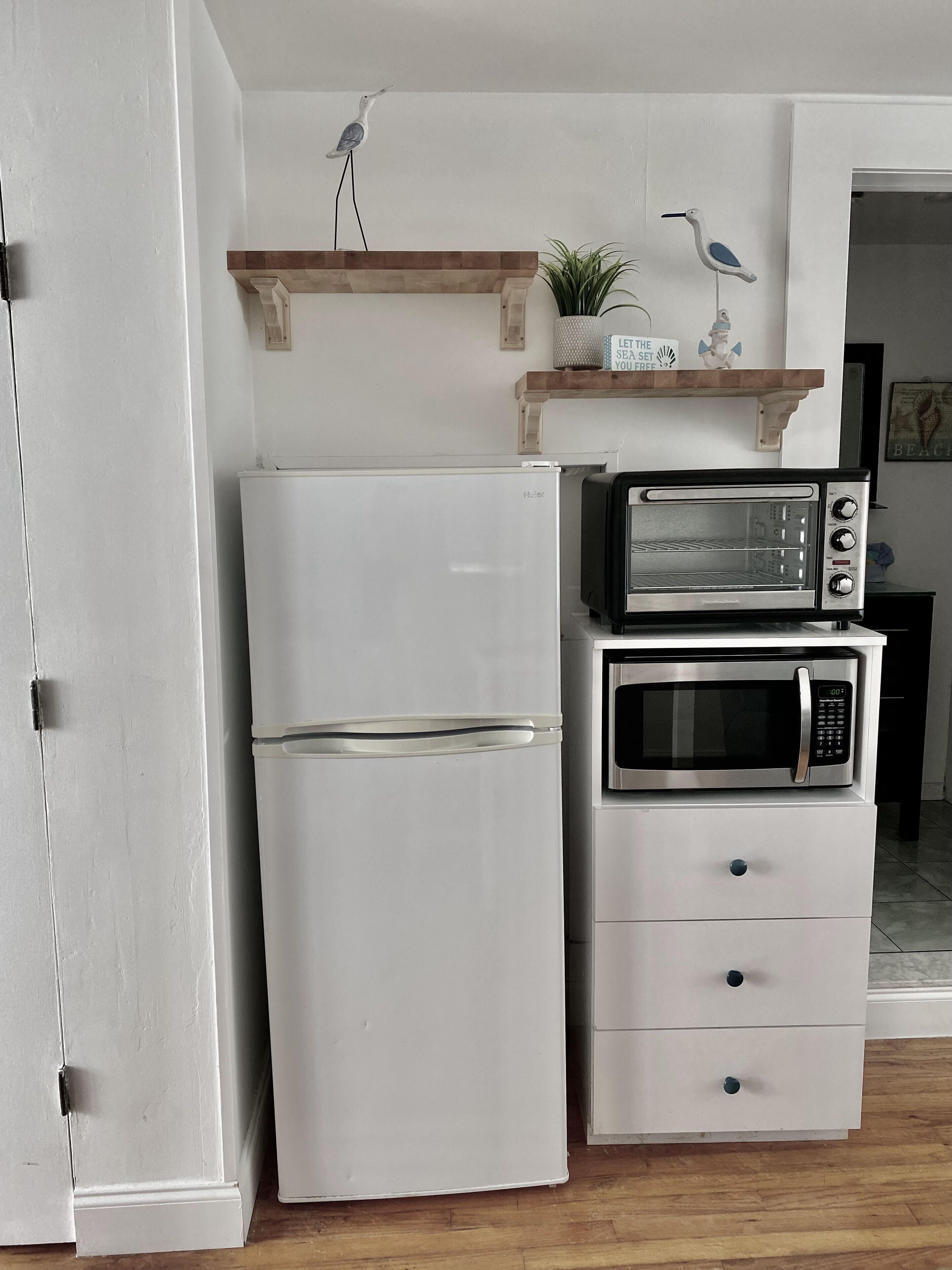 A compact kitchen with a white refrigerator, a microwave, and a toaster oven stacked on top of each other. There is a wooden shelf above the appliances with a small plant and a decorative item.