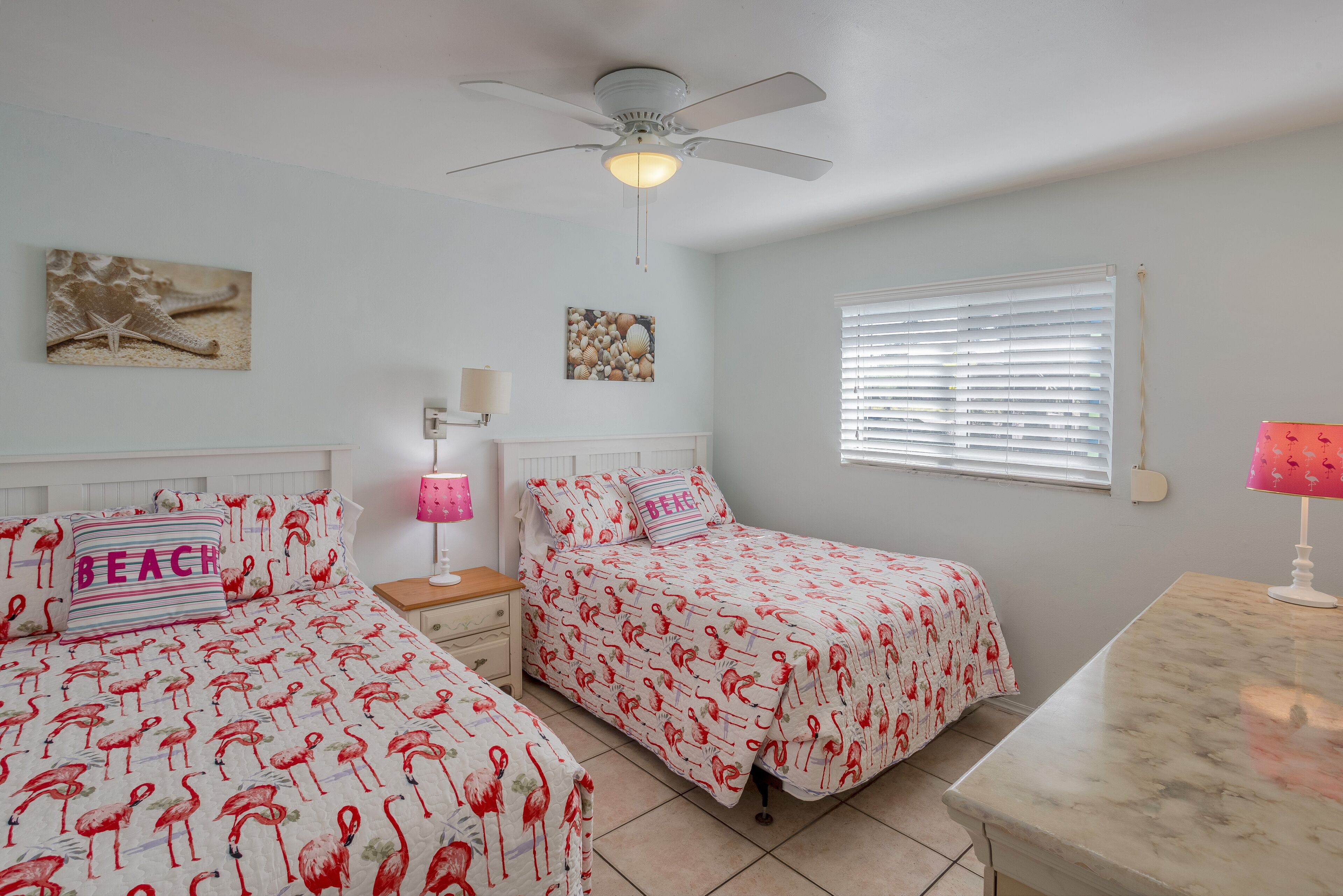 A bedroom with two beds, a ceiling fan, a window with blinds, and artwork on the walls.
