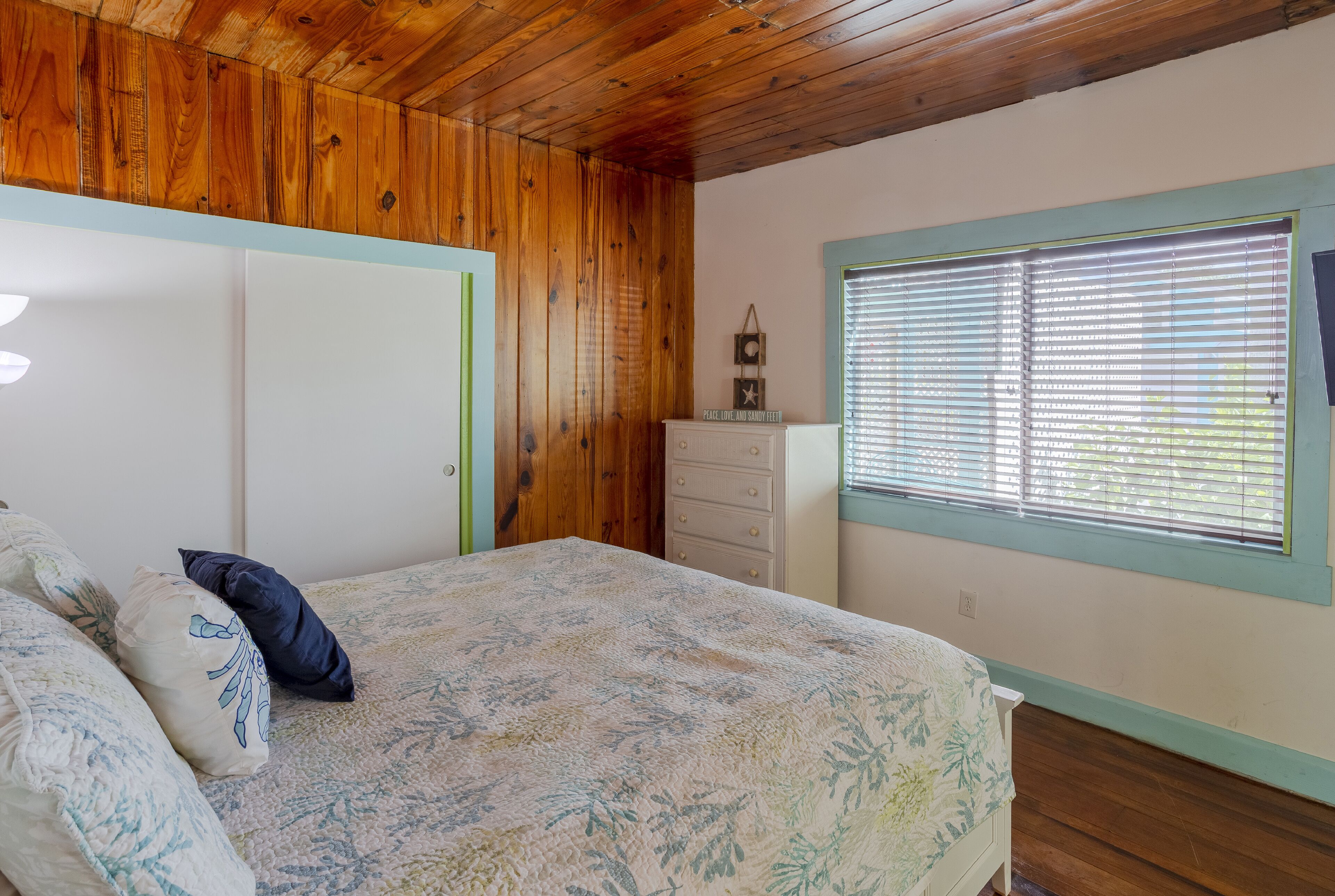 Un dormitorio con techo de madera, cama con ropa de cama floral, cómoda y ventana con persianas.