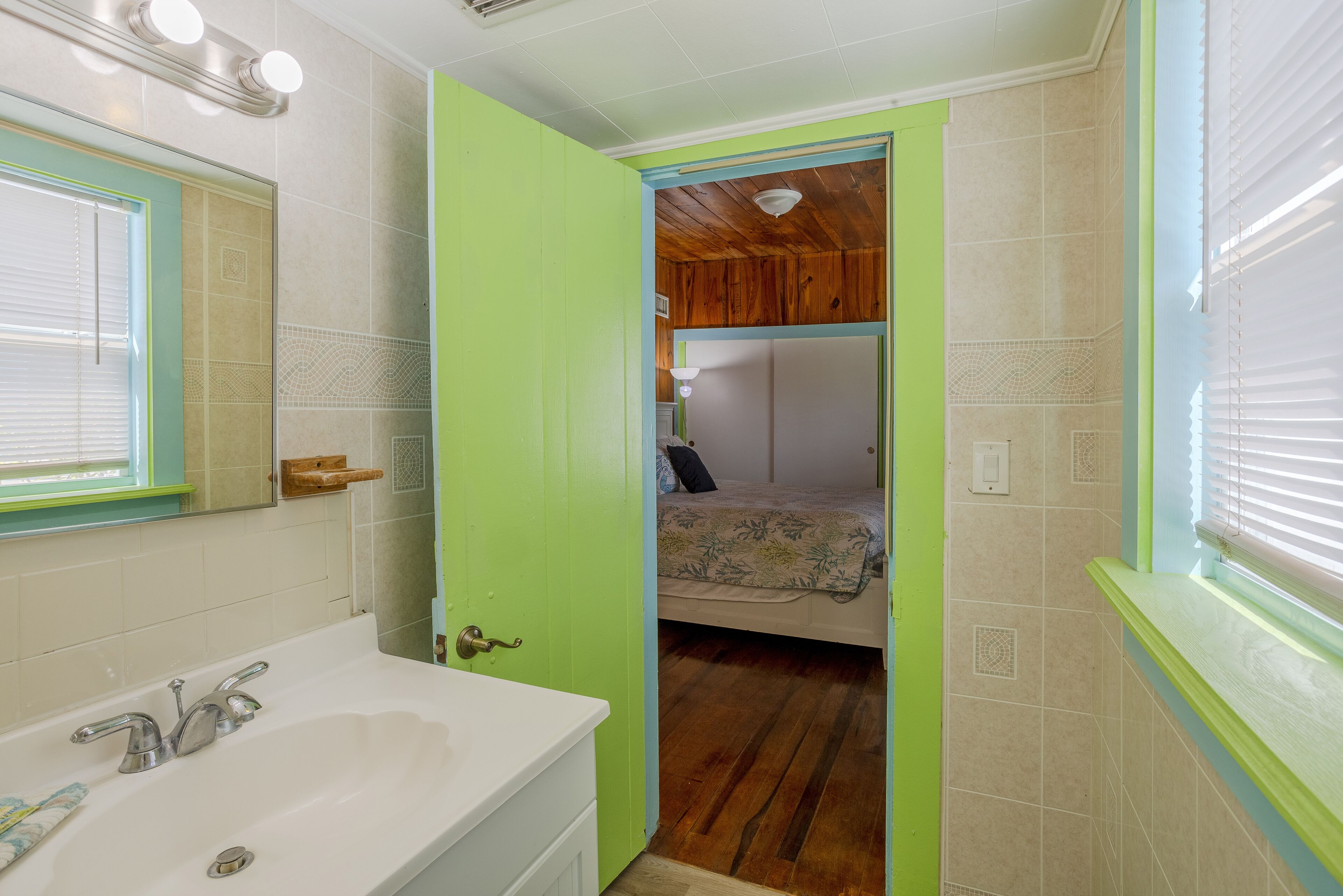 A bathroom with a white sink, a mirror, and a green door leading to a bedroom with a bed and wooden ceiling.