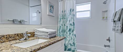 A bathroom with a granite countertop, a white sink, a toilet, a shower with a blue curtain, and a window.