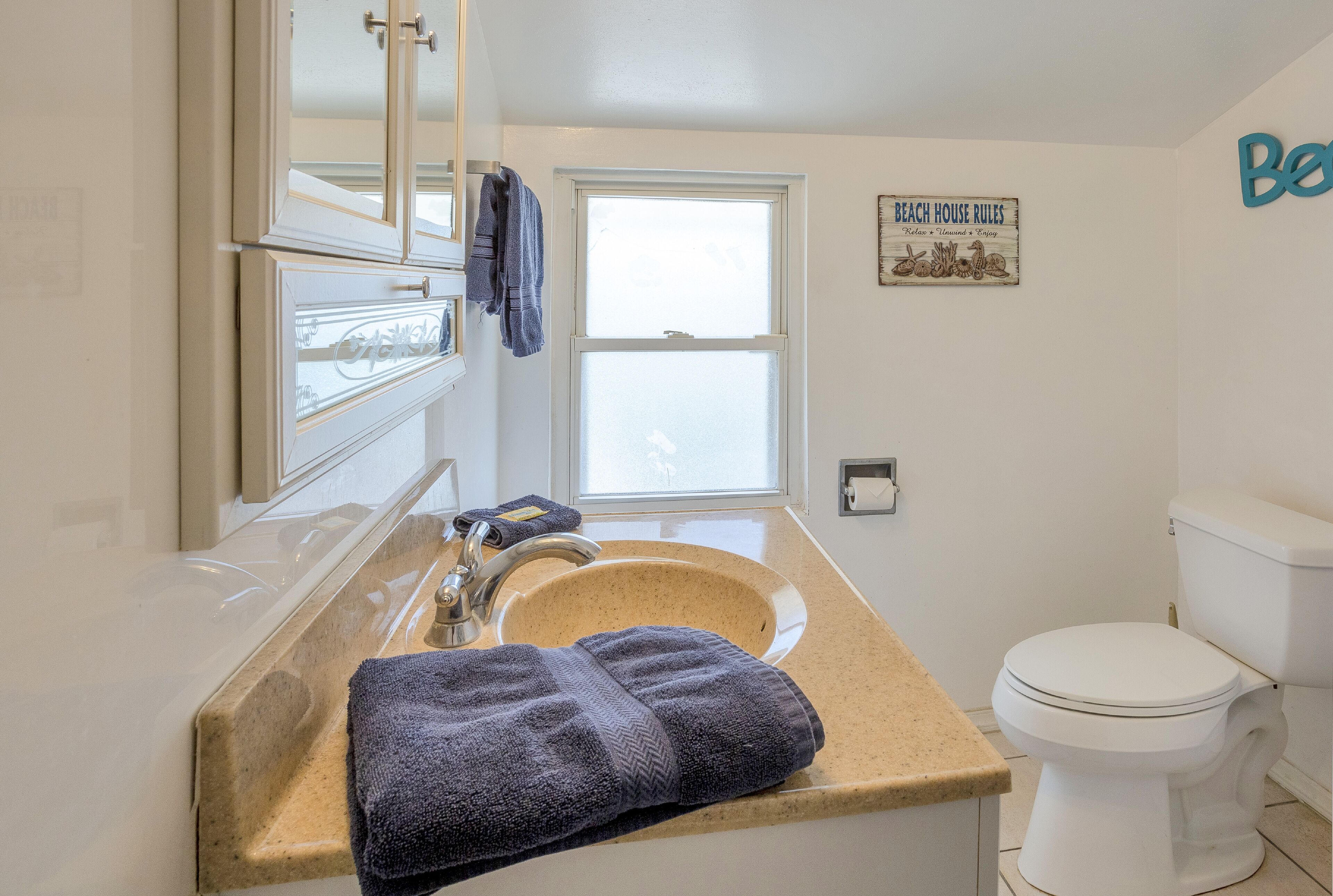 Un baño con encimera color beige, lavamanos blanco, espejo, toalla, inodoro y ventana.