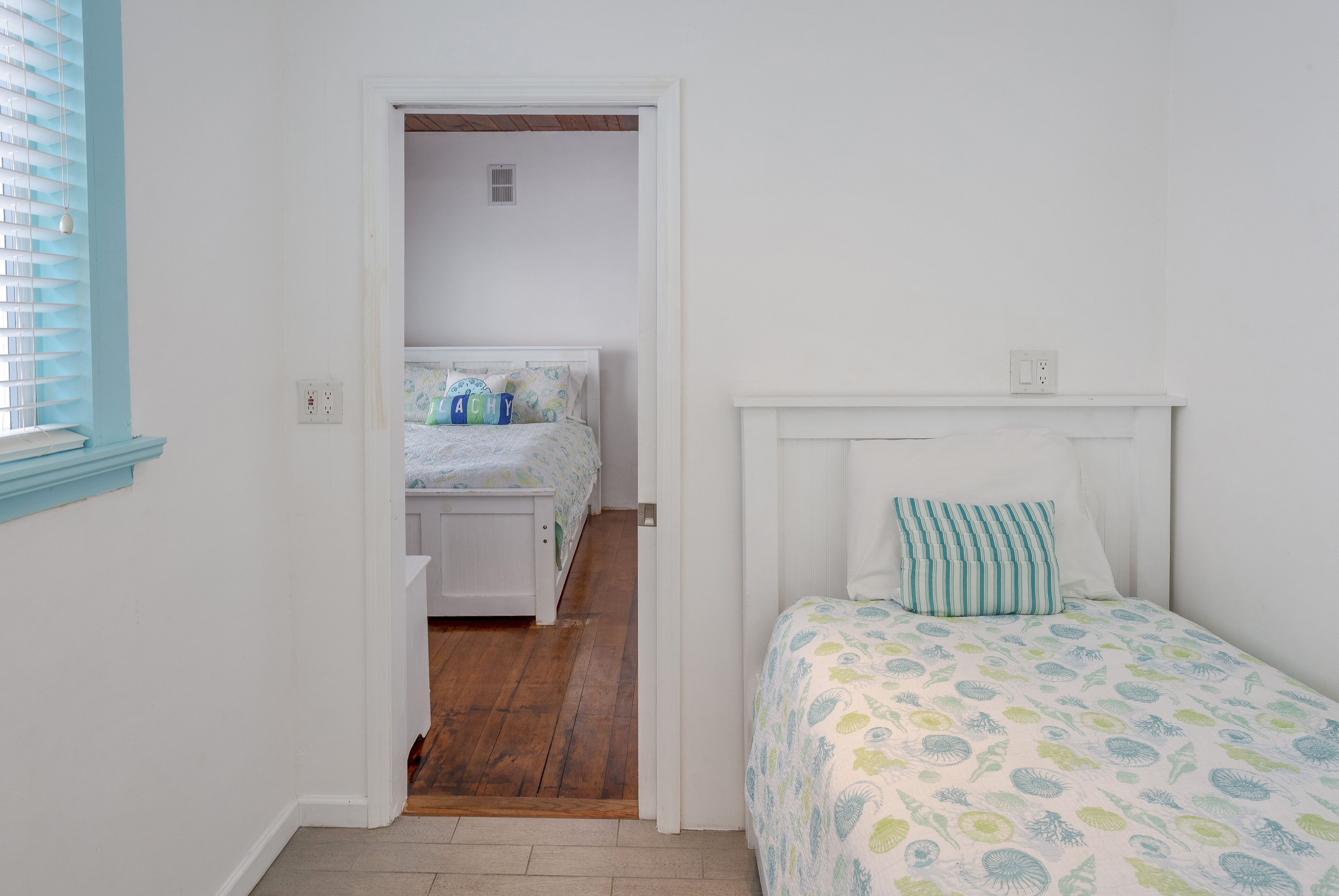 A bedroom with a wooden floor, white walls, a bed with a patterned bedspread, and a window with blinds.