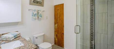 A bathroom with a granite countertop, a toilet, a glass shower enclosure, and a wooden door.
