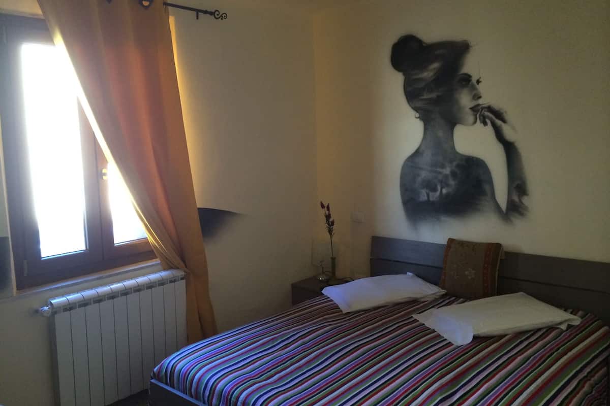 A hotel room with a bed, a radiator, a window with curtains, a wall-mounted artwork, and a bedside table with a lamp.