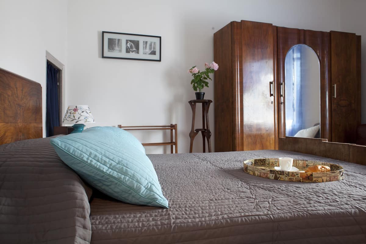 A bedroom with a bed, a wooden wardrobe, a small table with a plant, and a framed picture on the wall.