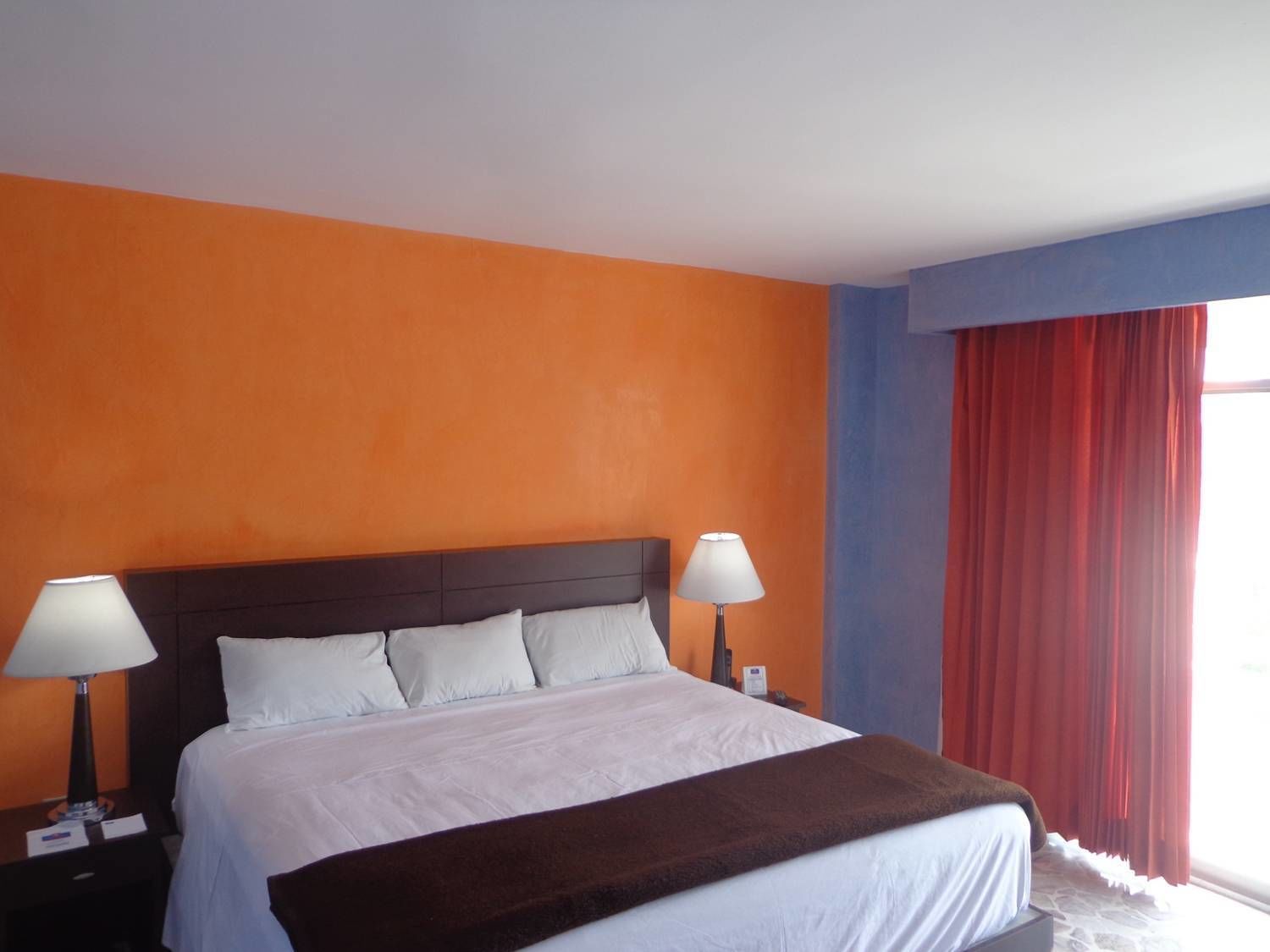 Habitación de hotel con pared naranja, una cama grande con ropa de cama blanca, dos lámparas de noche y una ventana con cortinas rojas.