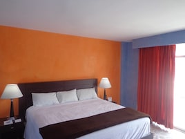 Habitación de hotel con pared naranja, una cama grande con ropa de cama blanca, dos lámparas de noche y una ventana con cortinas rojas.