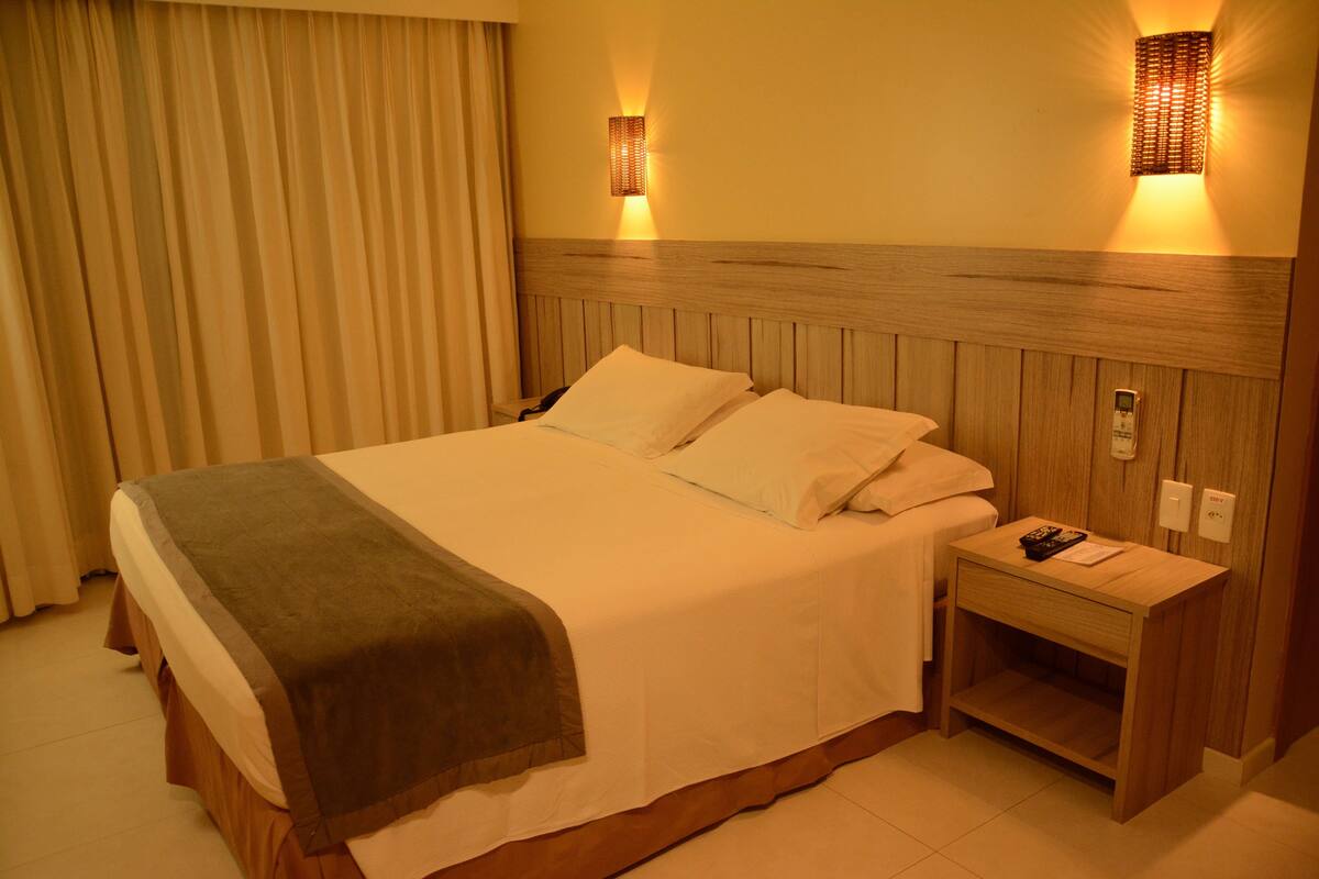 Une chambre d’hôtel avec un grand lit, une tête de lit en bois, deux lampes de chevet, une table de chevet et un rideau.