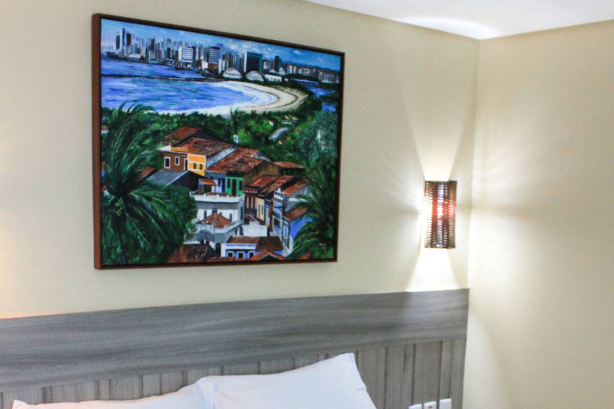 Un lit bien fait, avec des draps blancs et une couverture verte, un tableau représentant un paysage urbain côtier accroché au mur, et une lampe de chevet.