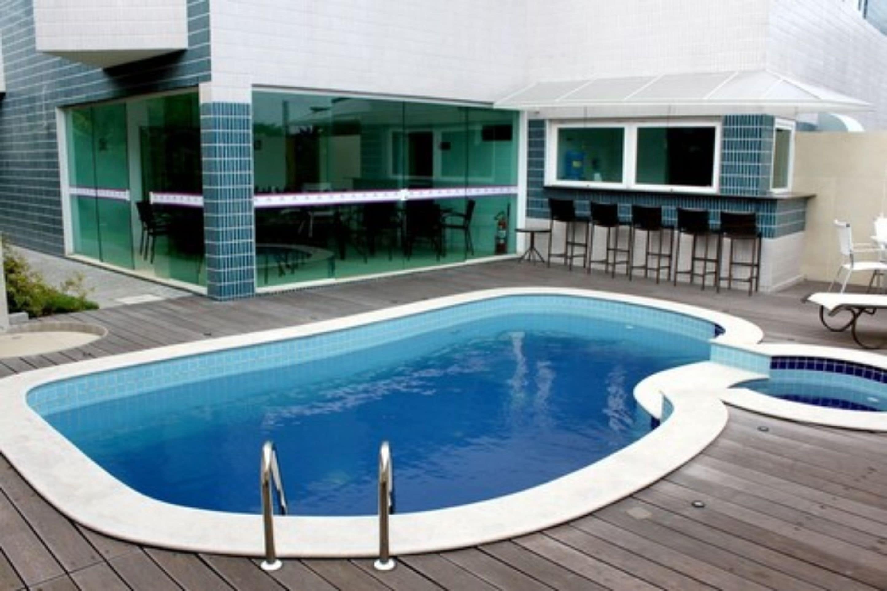 Una piscina exterior moderna con cubierta de madera, zona de bar con taburetes y puertas de cristal que dan a un espacio interior.