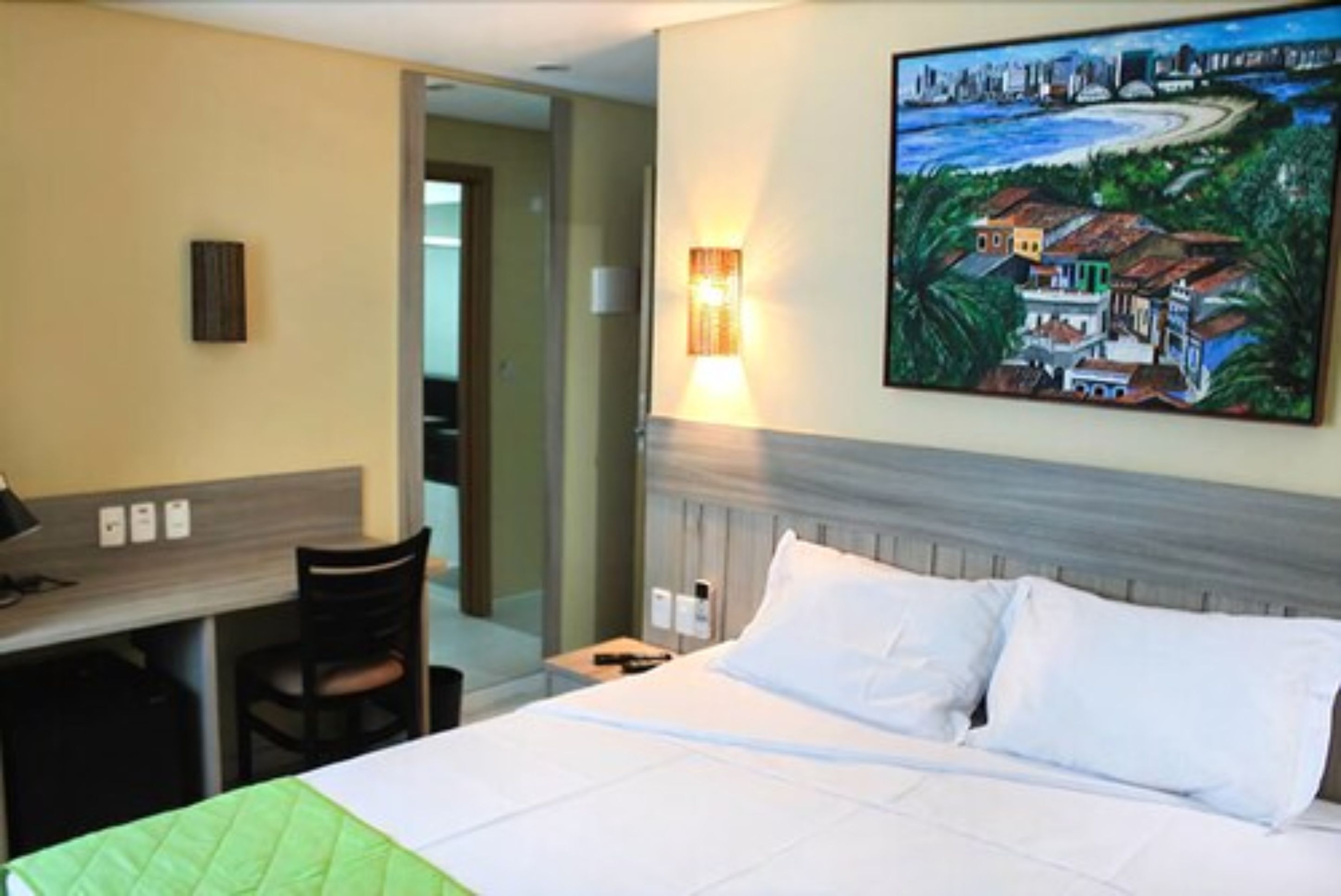 A hotel room with a bed, a desk, and a chair. There is a painting on the wall and a lamp on the desk.