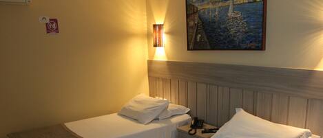 A hotel room with two single beds, a bedside table with a telephone and remote control, a wall-mounted light, and a painting above the beds.