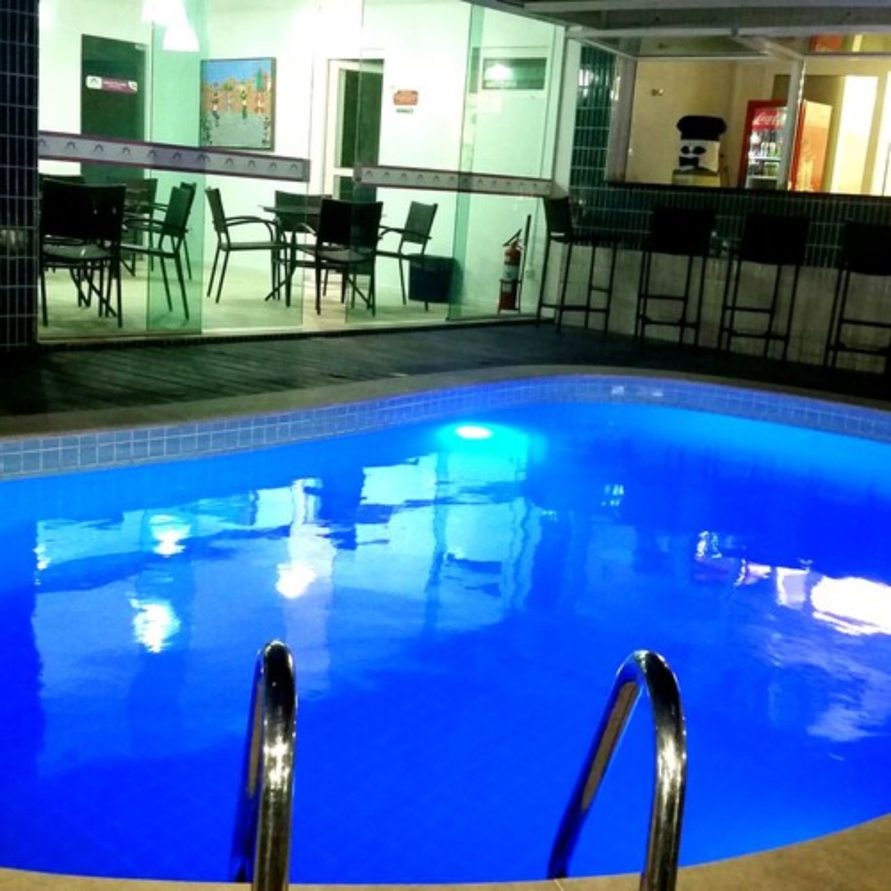 Una piscina con una escalera metálica, rodeada de sillas y mesas, y un refrigerador de Coca-Cola al fondo.