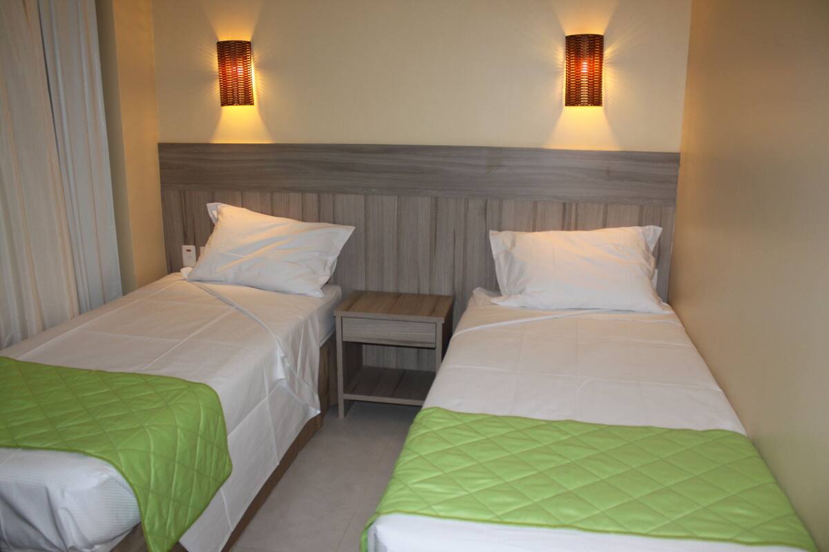 Une chambre d’hôtel avec deux lits simples, chacun recouvert d’une courtepointe matelassée verte et agrémenté d’oreillers blancs. Deux appliques murales sont fixées de part et d’autre de la tête de lit.