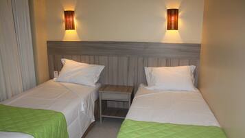 A hotel room with two single beds, each with a green quilted bedspread and white pillows. There are two wall-mounted light fixtures on either side of the headboard.
