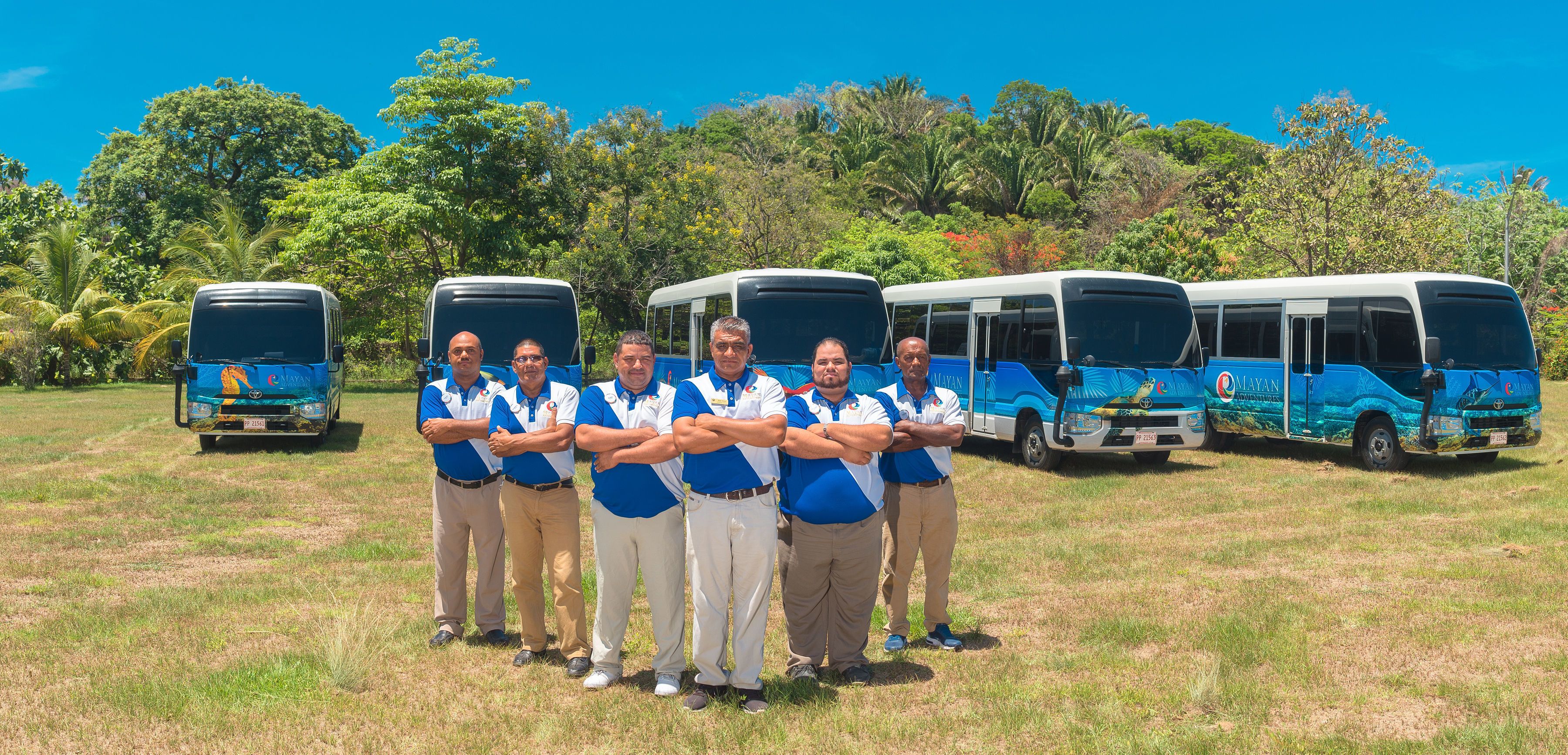A group of people standing in front of a row of buses with tropical trees in the background.