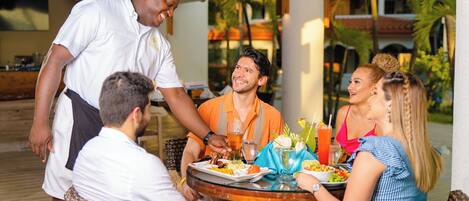 A group of people dining at an outdoor table with a waiter serving food.