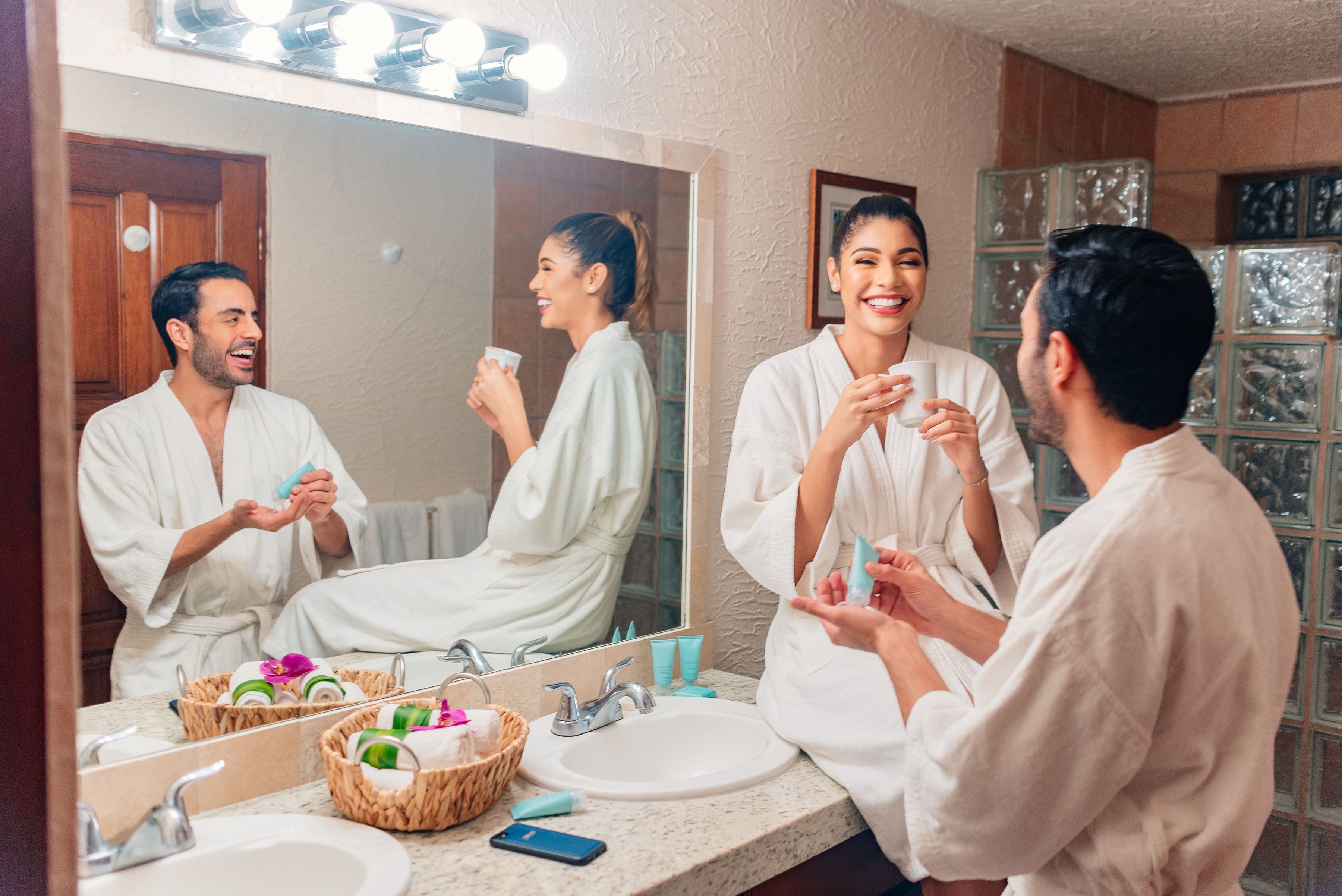 A bathroom with a large mirror, two sinks, a vanity, and a basket of toiletries.