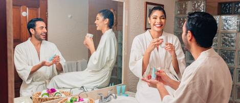 A bathroom with a large mirror, two sinks, a vanity, and a basket of toiletries.