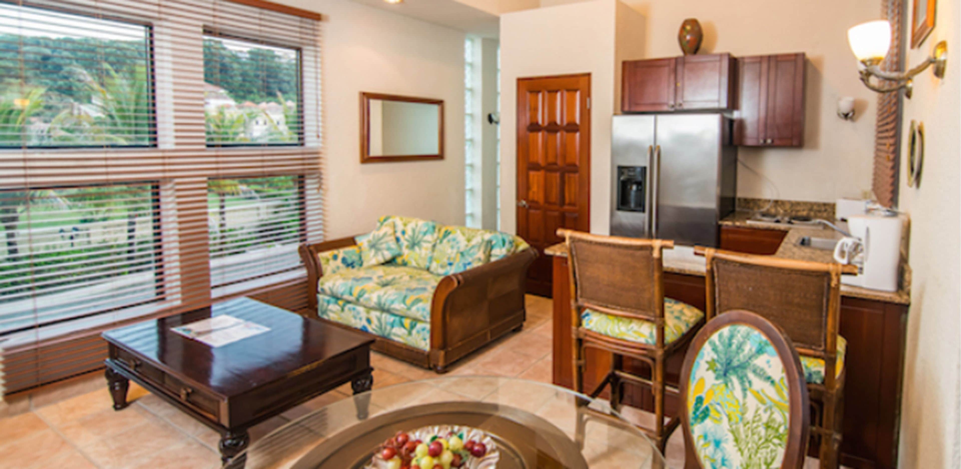 A living room with a sofa, a coffee table, and a dining area with chairs. There is a kitchen with a refrigerator and cabinets.