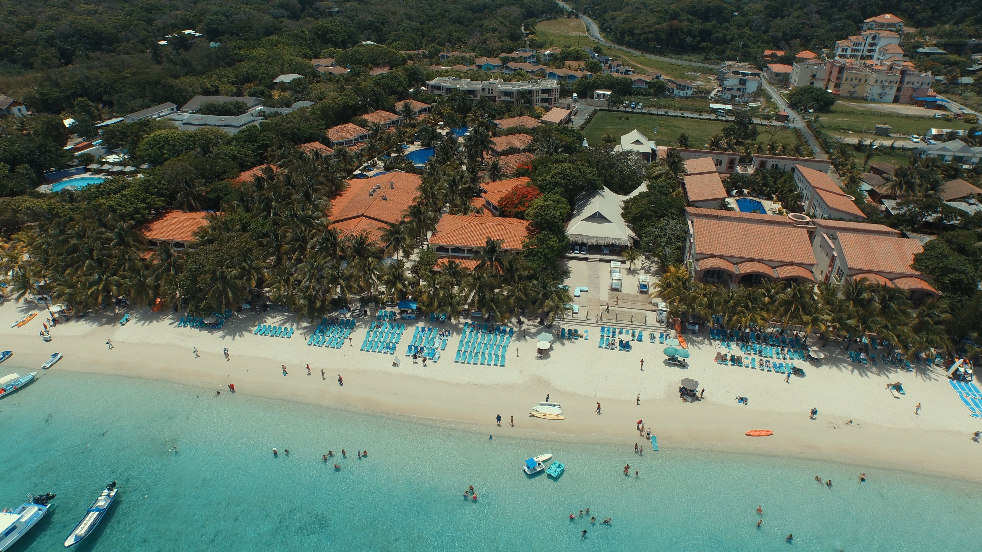 An aerial view of a coastal resort with a beach, multiple buildings with red roofs, and a pool area.