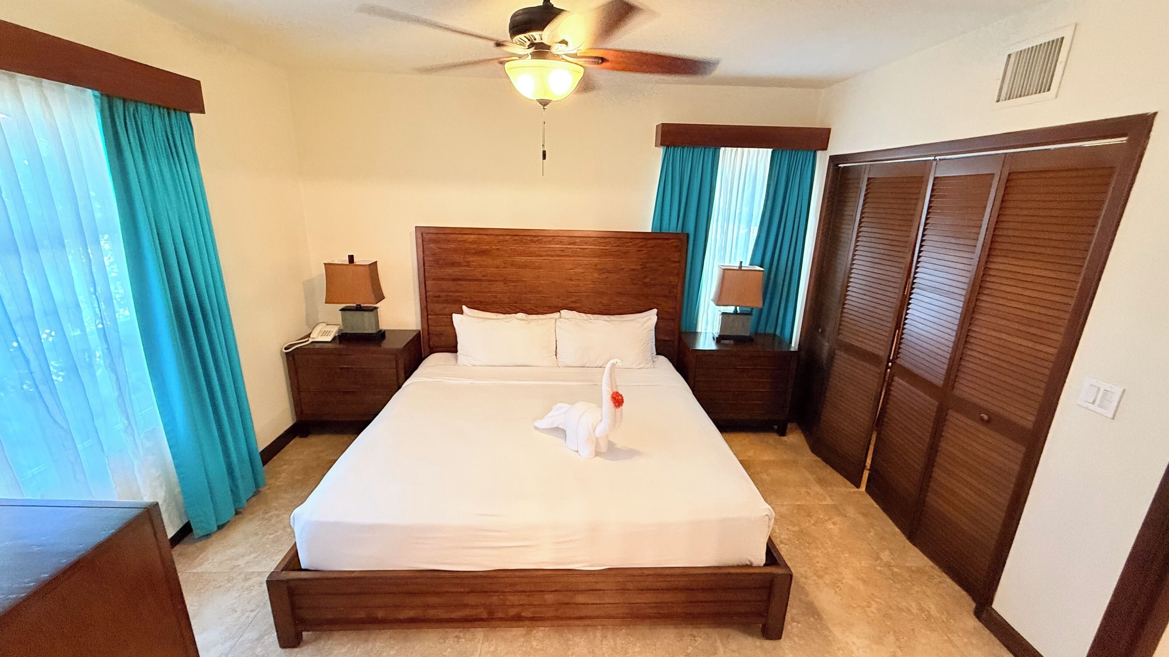 Habitación de hotel con cama de madera, dos mesitas de noche con lámparas, ventilador de techo y armario.