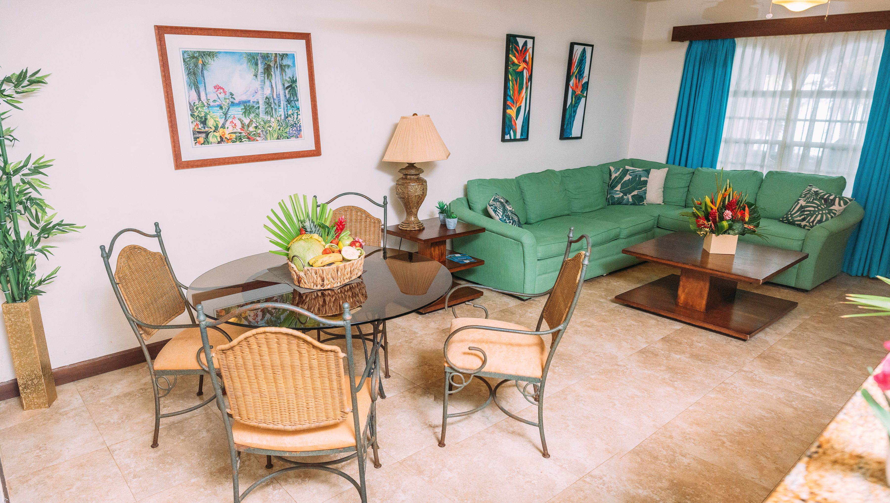 Una sala de estar con un sofá verde, una mesa redonda de vidrio con sillas, una lámpara y un cuadro enmarcado con motivos tropicales en la pared.