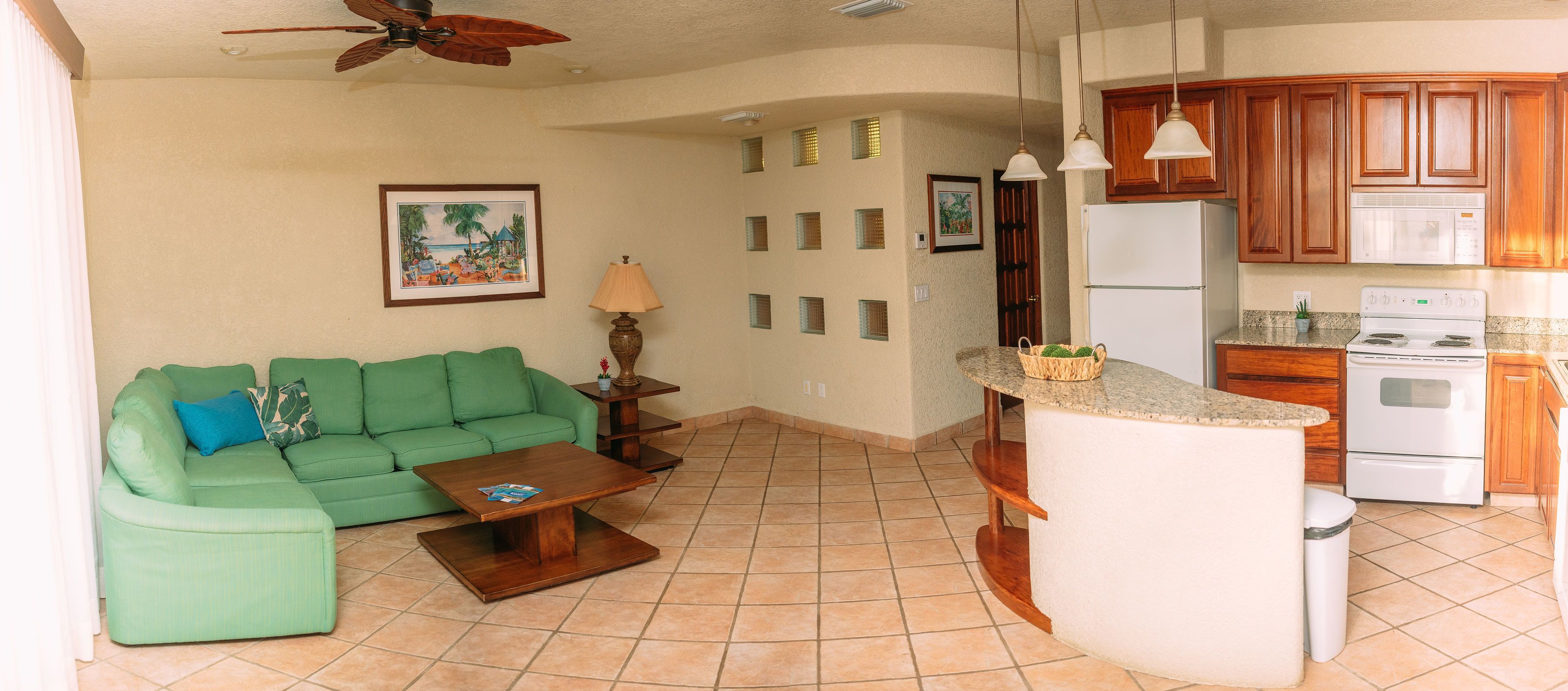 A living room with a green sofa, a coffee table, and a kitchen area with wooden cabinets and a white refrigerator.