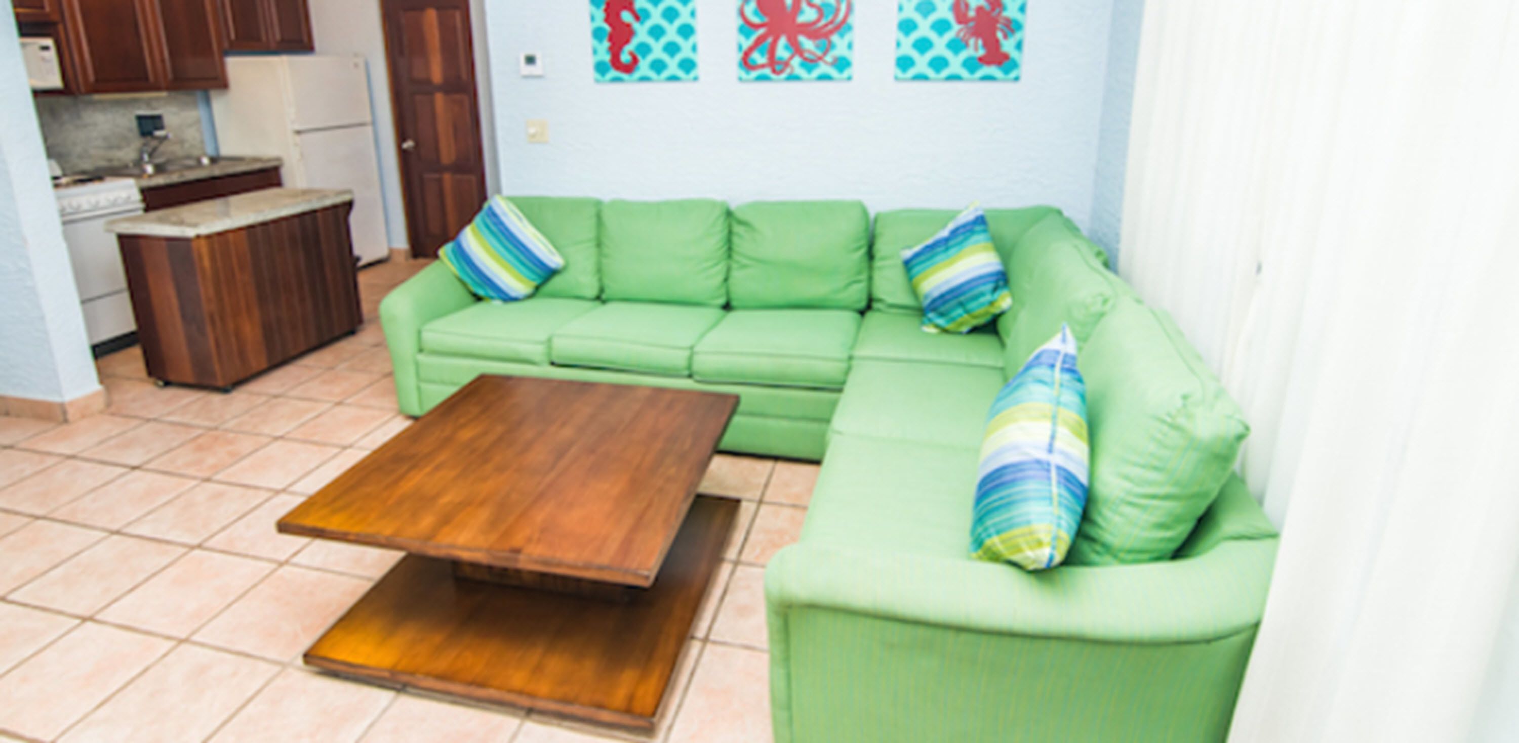 Un sofá verde con almohadones a rayas, una mesa de centro de madera y una cocina con armarios de madera y un refrigerador blanco.