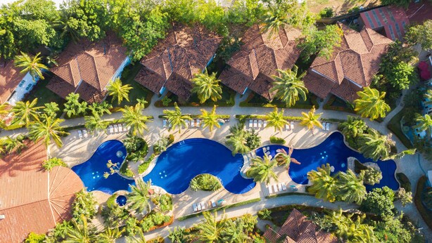 An aerial view of a resort with multiple bungalow-style buildings, a central swimming pool, and surrounding palm trees. - Mayan Princess Beach & Dive Resort - All Inclusive (Roatan)