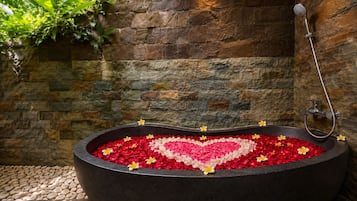 A bathtub filled with rose petals and a heart-shaped arrangement, with a showerhead and stone wall backdrop.