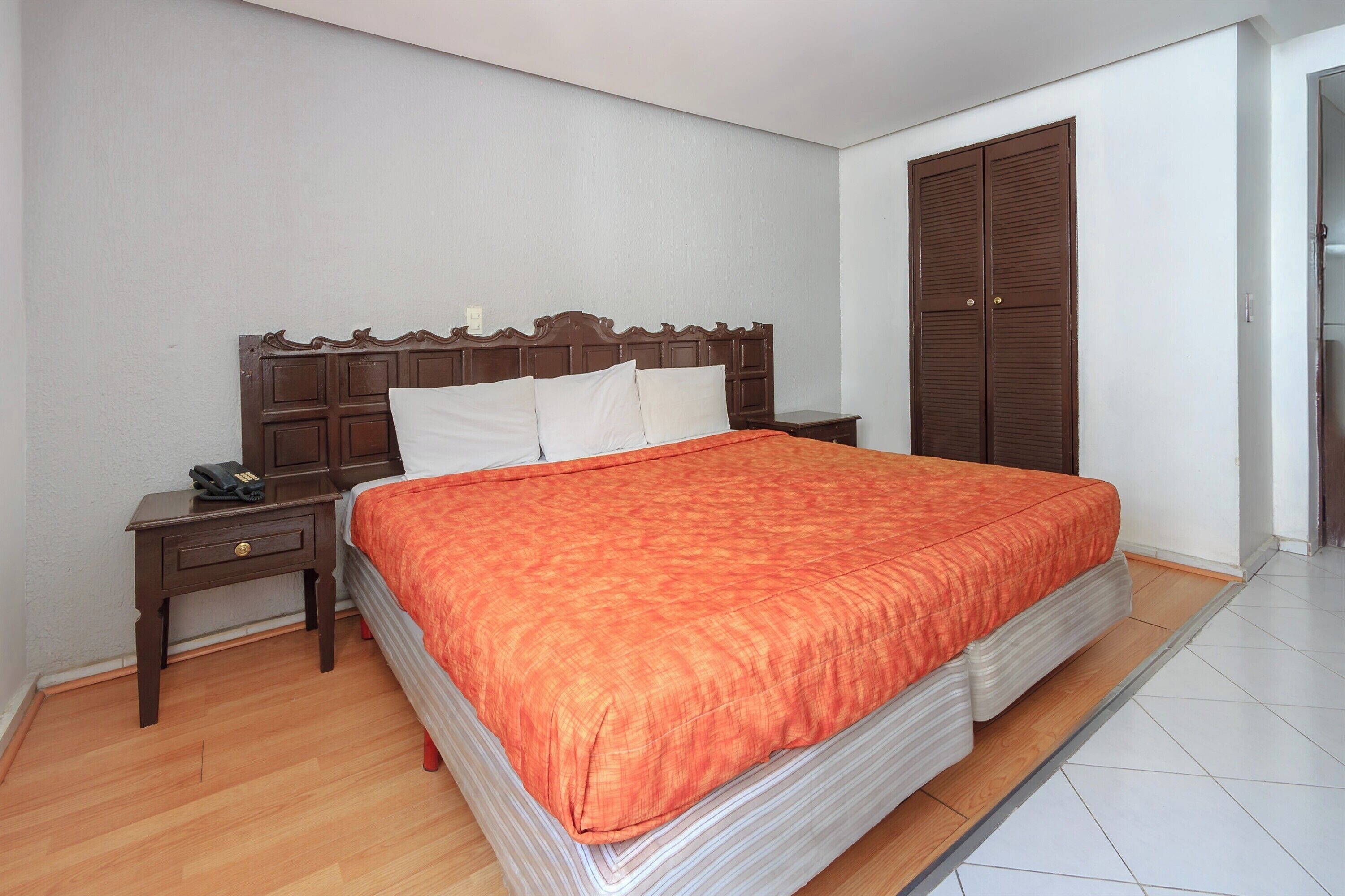 A bedroom with a large bed, wooden headboard, white pillows, an orange bedspread, a nightstand with a phone, and a wooden wardrobe.