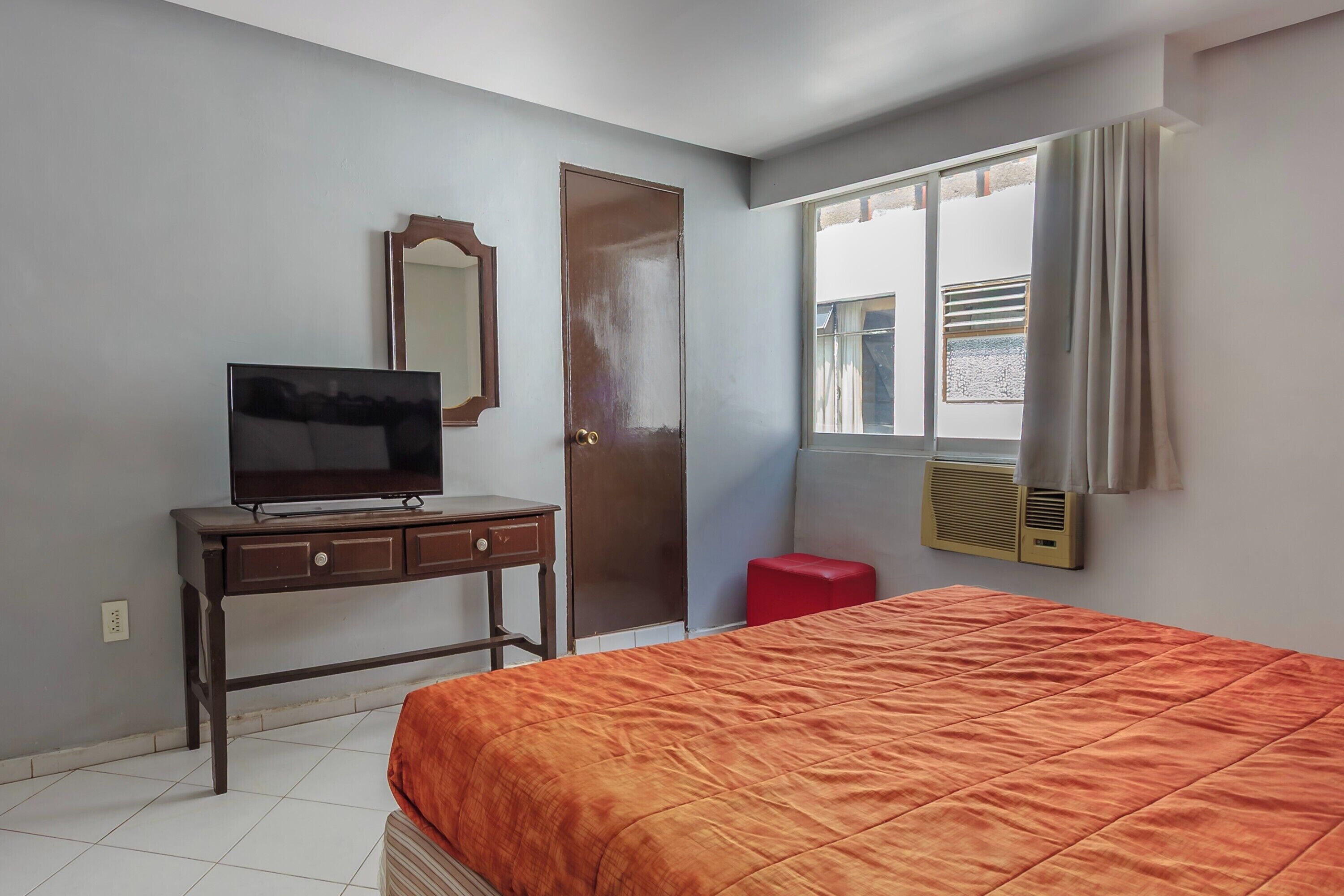 A bedroom with a bed, a TV on a desk, a door, and a window with curtains.