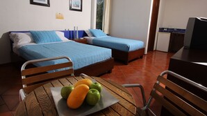 A bedroom with two beds, a wooden table with fruit, and a kitchenette in the background.