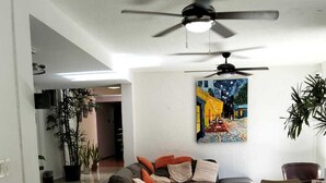 A modern living room with a sofa, dining table, chairs, ceiling fan, and a painting on the wall.