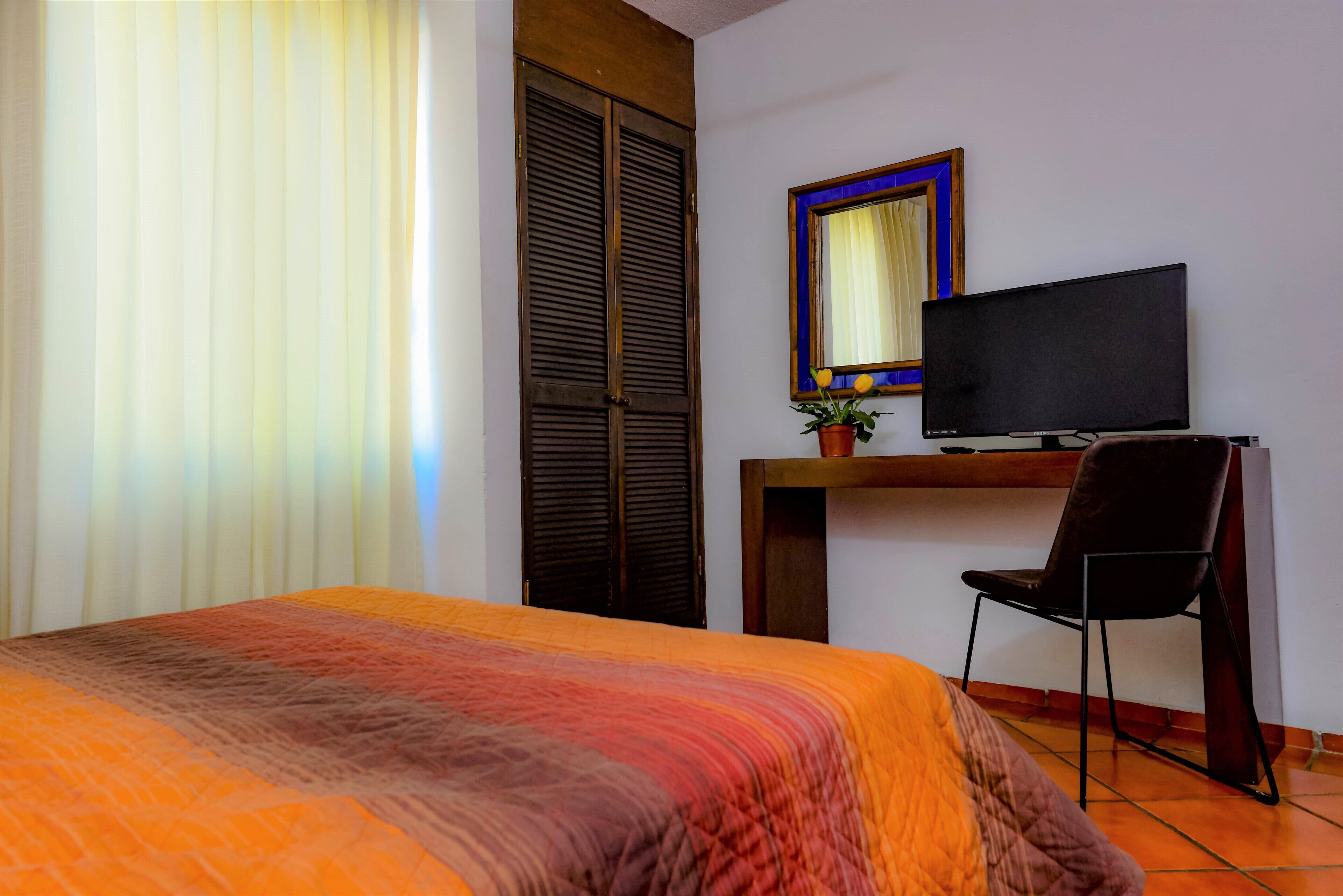 Un dormitorio con una cama, un televisor sobre un escritorio, una silla y una ventana con persianas.