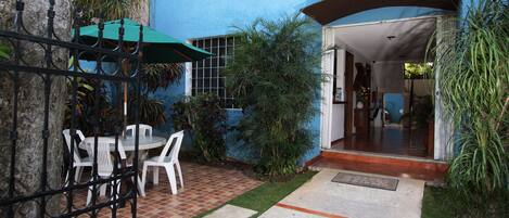 Un patio con suelo de baldosas, sillas blancas, un paraguas verde y una reja de metal.
