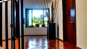 A hallway with tiled flooring, a window with a view of greenery, and a black speaker in a black stand.