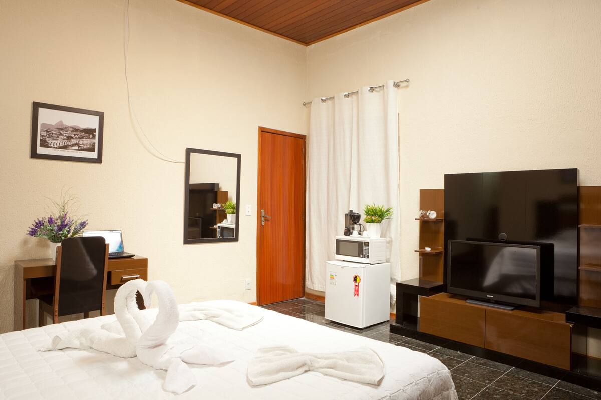 A hotel room with a bed, a desk with a laptop, a microwave, a TV, and a door with a curtain.