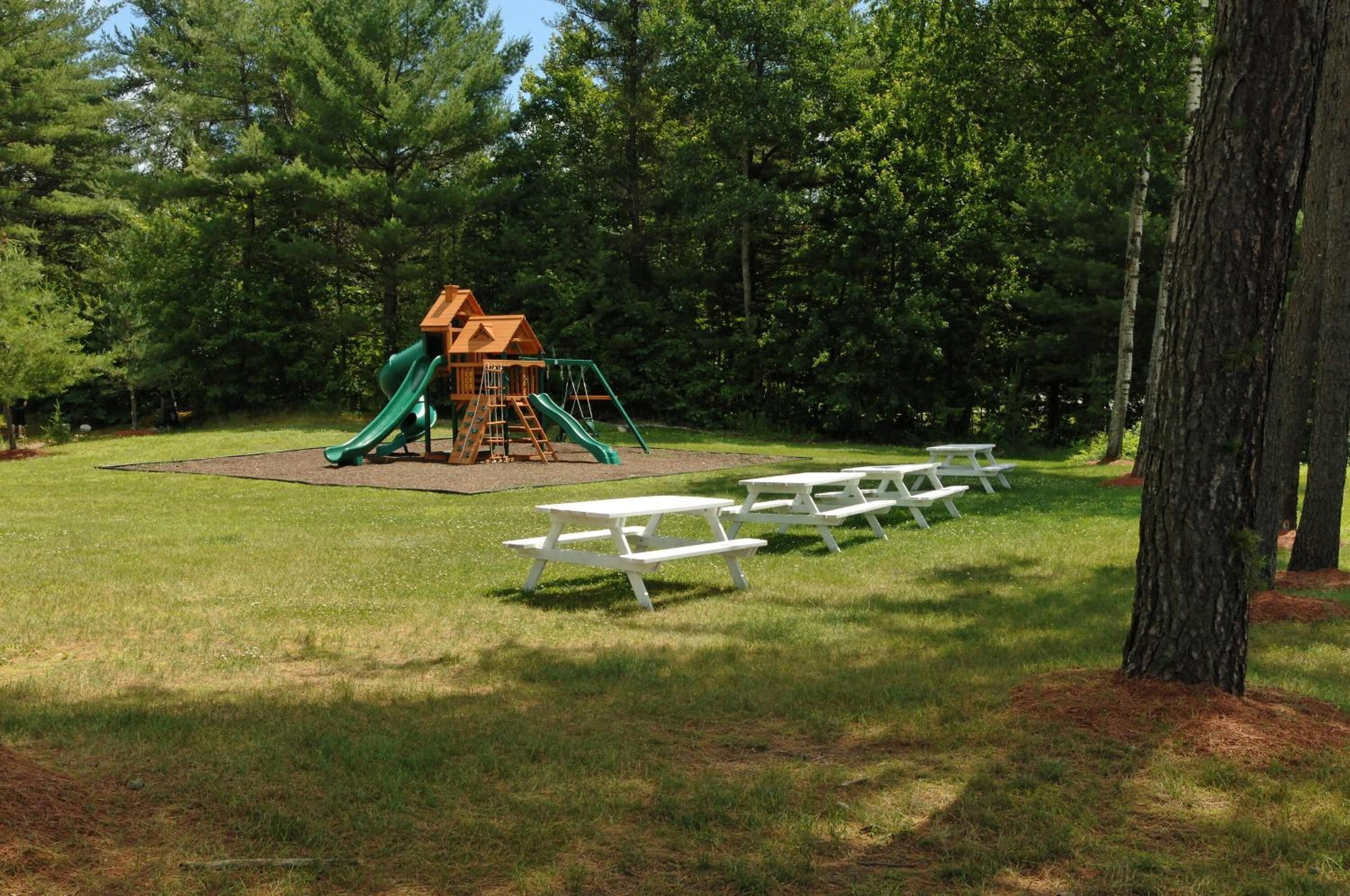 A playground with a wooden playhouse and slides, surrounded by picnic tables and trees.